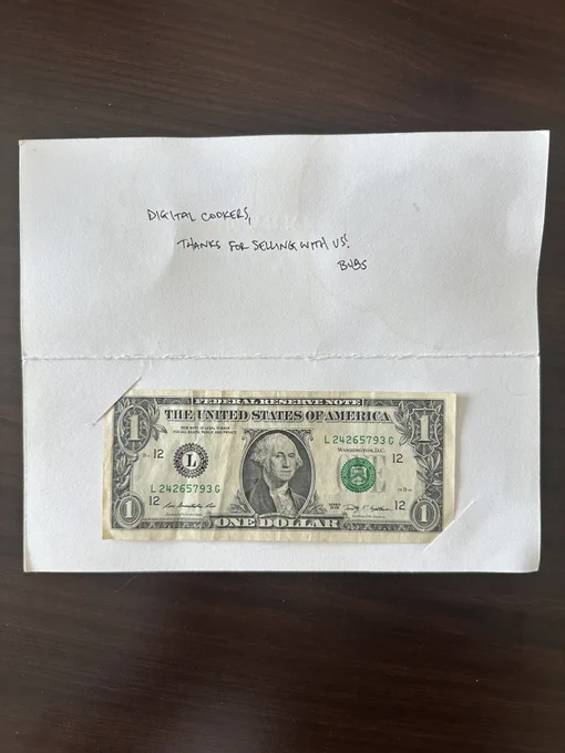 A handwritten note on a folded white card reading "Digital Cookers, thanks for selling with us! Bubs," with a one-dollar bill taped below it, resting on a dark wood desk.
