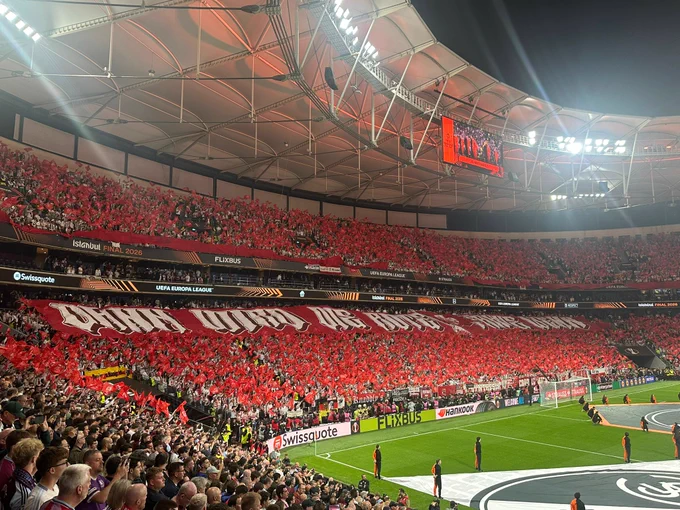 Crowded football stadium with supporters in white and red holding up a massive banner reading “OUR TIME IS NOW” before a UEFA Europa League match.