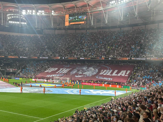 Packed football stadium before kick-off with thousands of supporters displaying a large claret and blue tifo reading “UP THE VILLA!” during a UEFA Europa League match.