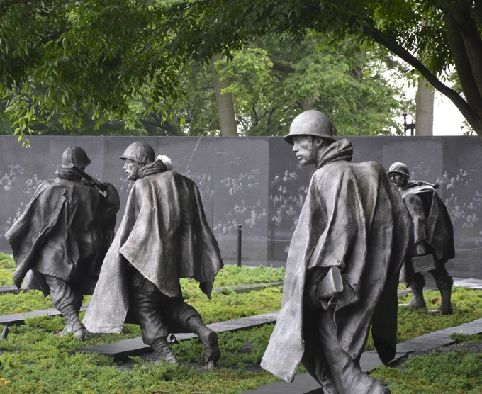 Four large gray metals statues of soldiers wearing ponchos and helmets, walking in formation at the Korean War Veterans Memorial surrounded by greenery.