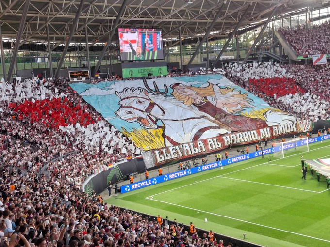 Stadium grandstand filled with fans waving red and white flags around a massive central banner illustrating a woman riding a white horse. A banner below reads "LLÉVALA AL BARRIO, MI AMOR."