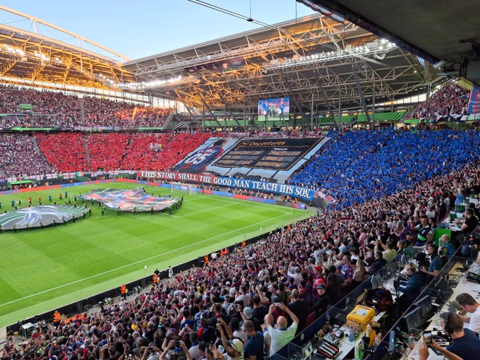 A packed stadium with stands split into red and blue fan sections. A massive airport departures-themed tifo banner hangs in the center above a banner reading "THIS STORY SHALL THE GOOD MAN TEACH HIS SON."