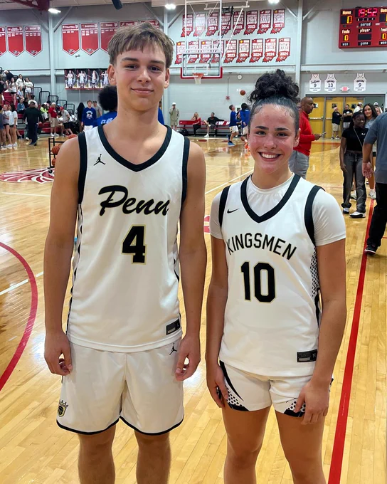 Two basketball players in white jerseys, one boy and one girl, standing on a basketball court with scoreboard and other players in the background.