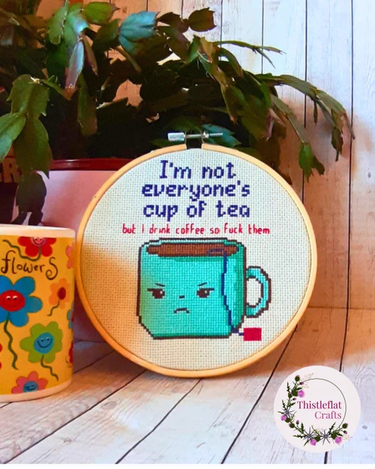 Funny cross stitch in wooden hoop reading 'I'm not everyone's cup of tea. But  I drink coffee so fuck them' featuring cute but grumpy cup of tea, styled with and plant and a mug on a rustic wooden background – handmade sarcastic embroidery wall art