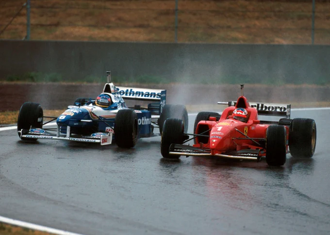 Both cars kicking up spray, the blue and white Rothmans sponsored Williams FW18 of Jacques Villeneuve is on the left of frame, being passed on the inside by the red Marlboro sponsored Ferrari F310 of Michael Schumacher, on the right of frame.