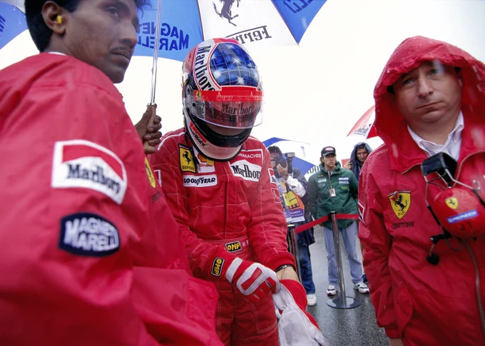 In the centre, Michael Schumacher stands in his red race suit and blue topped white crash helmet with the visor open. He is looking down as he pulls his race gloves on. On the left of frame, Balbir Singh stands holding a blue and white Ferrari branded umbrella over Schumacher. On the right of frame, Jean Todt stands wearing a red rain jacket with the hood up, headphones tucked into the front.