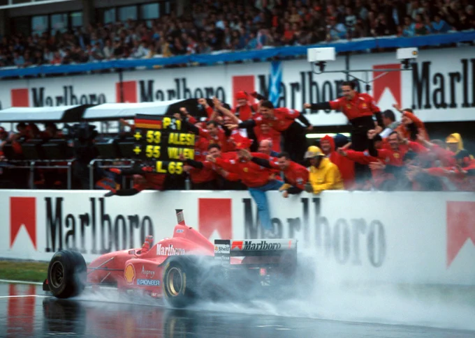 Michael Schumacher's red Ferrari F310 kicks up a large amount of spray as it passes the Marlboro branded pit wall. A number of Ferrari team members hang over the low pit wall, punching the air or waving, with one member holding out a pit board indicating Schumacher's huge lead.