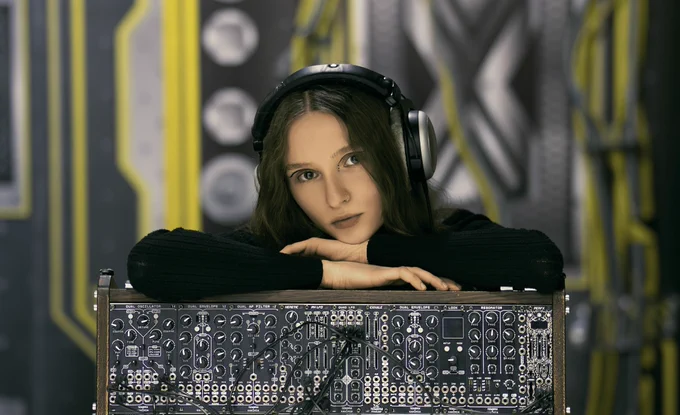 Kaximia, wearing headphones, rests her head on the top of a skiff of Eurorack gear from Ukrainian maker Tembra Modular.