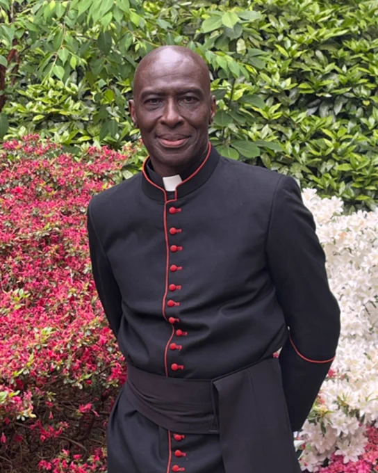 A person in formal clerical attire stands outdoors among vibrant flowering bushes and lush greenery, smiling at the camera.
