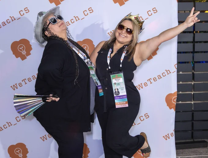 Two women wearing shades and facing each other pose for a photo. One woman throws up a peace sign while the other tilts her head back with a hand on her hip.