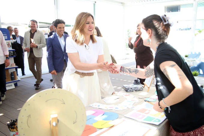 La alcaldes estrecha la mano de una educadora ambiental frente a una mesa con folletos y una rueda de colores en un evento.