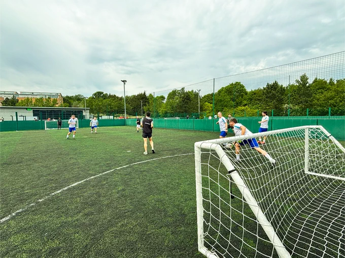 A group of people playing a football match on a green turf field, with one person kicking the ball toward a goal. Others on the field are focused on the action. Trees and a building are visible in the background under a cloudy sky.