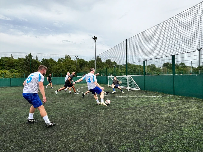A group of people play soccer on an outdoor field. One player, wearing a jersey with the number 4, is kicking the ball towards the goal, where a goalkeeper is ready to defend. Other players look on. The field is surrounded by tall green fences and trees in the background.