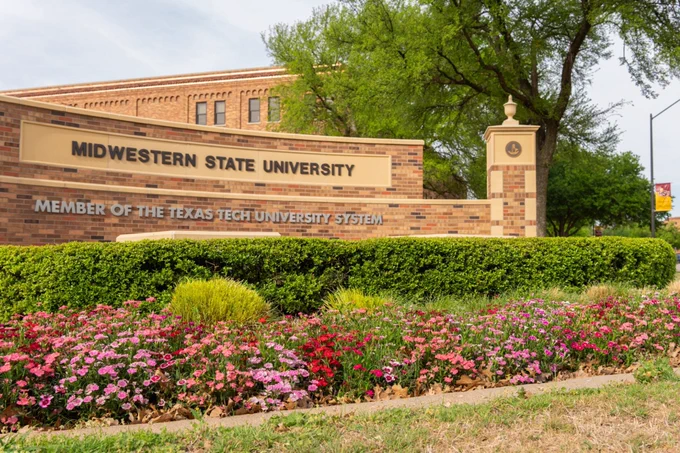 Exterior view of the Midwestern State University entrance sign featuring brick architecture, colorful flower beds in the foreground, and green trees surrounding the campus entrance on an overcast day. The sign reads “Midwestern State University” and “Member of the Texas Tech University System.”