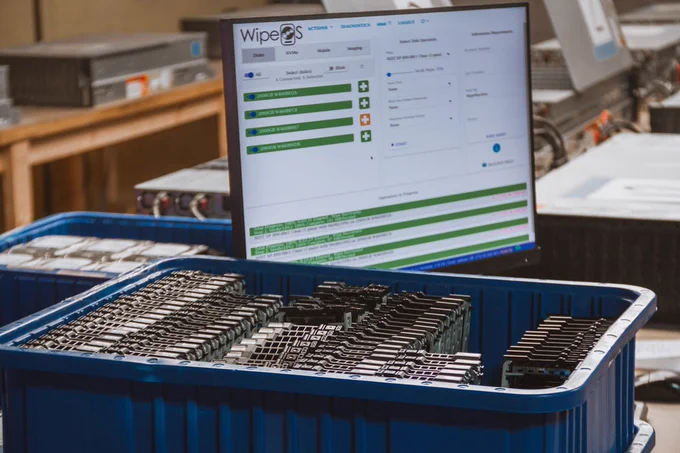 A close-up of a rugged laptop in a server aisle showing a detailed data sanitization dashboard with technical drive specifications and status logs.