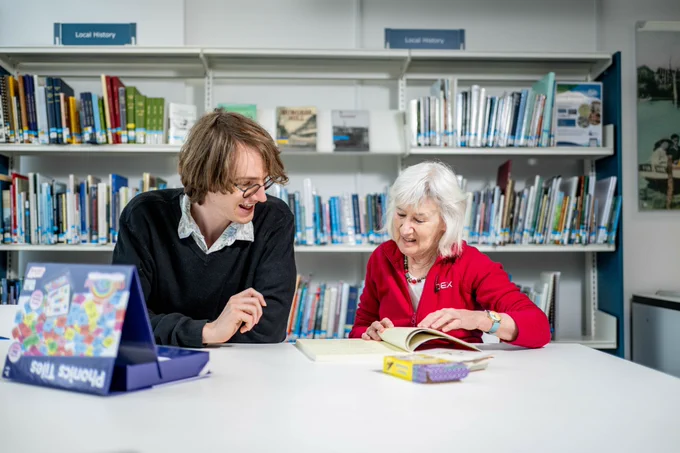 A young man and an older woman are sitting at a table in a library, looking at a book together.