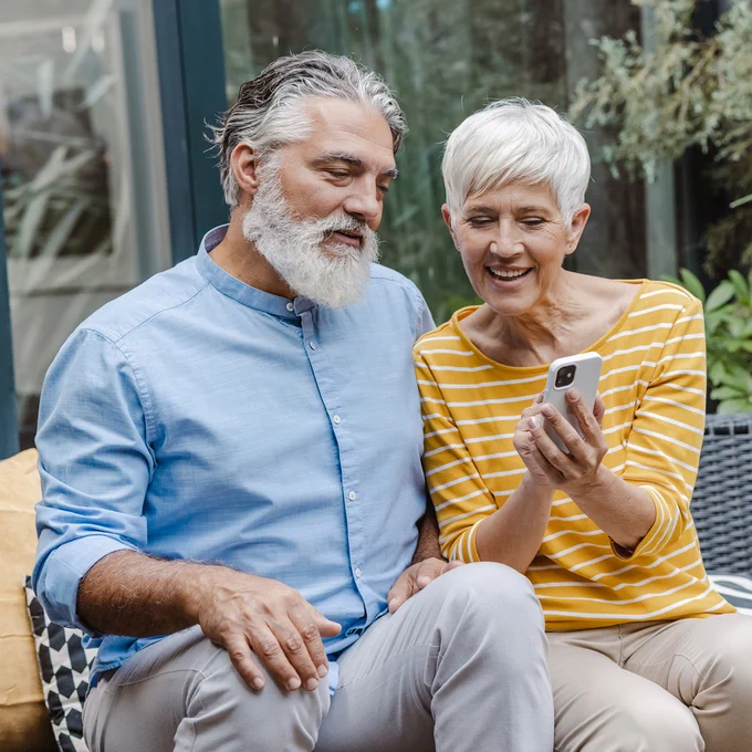 A senior couple sitting together outdoors looking at a smartphone.
	
