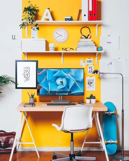 Desk setup with a yellow accent wall, shelves hold decor, including a clock and plants. A computer displays a floral image. A framed number art \"123\\n456\\n789\" is on the desk, with 'u.', 'coffee...', 'Sunday', 'YEAH', and 'enjoy' texts scattered.