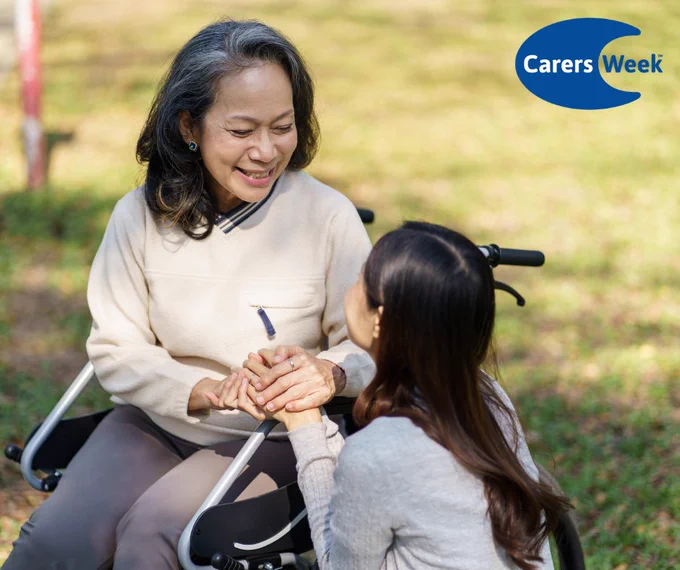 A woman kneeling outdoors holds hands with an elderly person seated in a wheelchair during a sunny day.