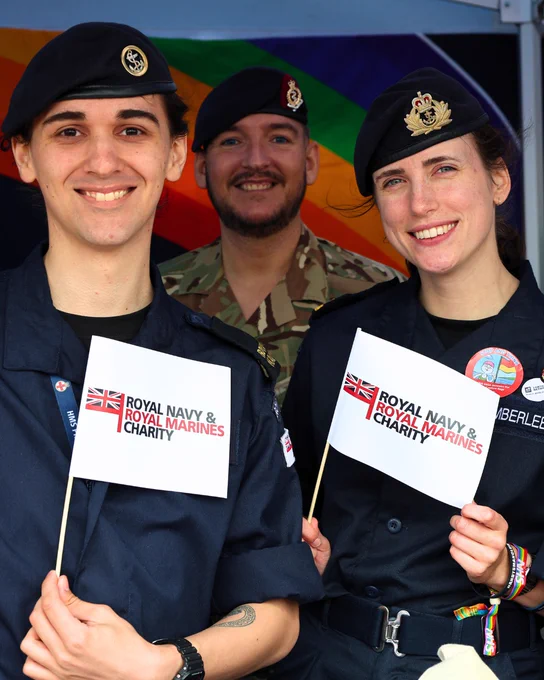 A group of three individuals in military uniforms smiling and holding small flags that read "Royal Navy & Royal Marines Charity." A colorful rainbow flag is visible in the background.