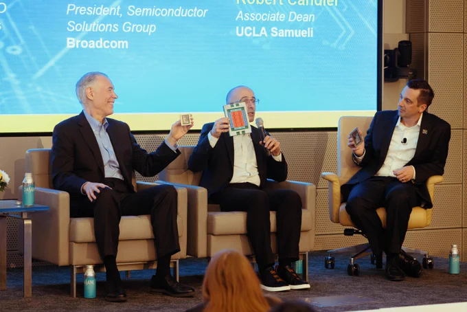 Three individuals are seated on a stage, engaged in a panel discussion. The person on the left holds up a small electronic component. The person in the center shows a colorful diagram. The background features a screen displaying names and affiliations, including Broadcom and UCLA Samueli.