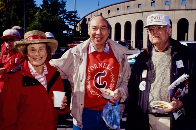 A group of three people is enjoying an outdoor event. They are wearing clothing with "Cornell" logos, suggesting a connection to Cornell University. The background shows a stadium, indicating a possible sports or university-related event. One person is holding a drink, while the others have plates of food. They are all smiling, creating a cheerful atmosphere.