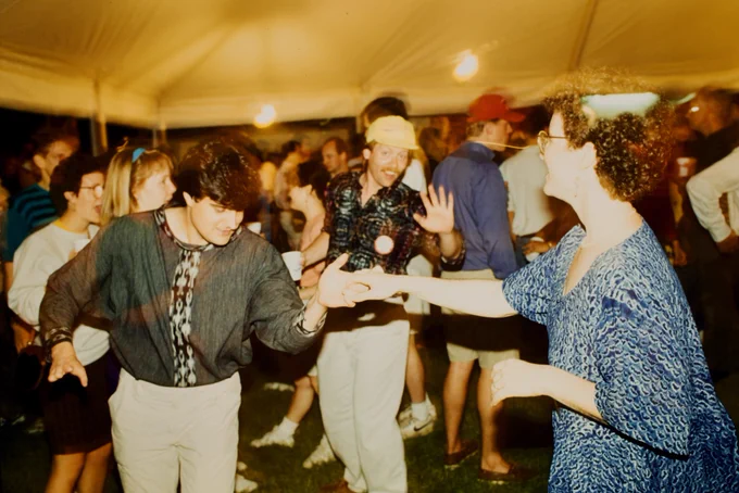 A lively group of Cornell reuniongoers dancing under a large tent on the Cornell Arts Quad. Some individuals are holding hands and smiling, while others chat in the background. Most are casually dressed.