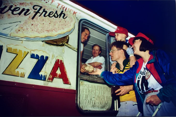 A group of people are gathered at a red and white food truck with "Oven Fresh Pizza" painted on the side. One person inside the truck is handing something to those outside. The scene is lively and is part of an undated Cornell Reunion.