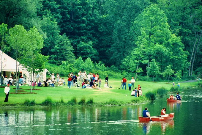 A lively outdoor scene by Cornell's Beebe Lake at an undated Cornell Reunion. People are gathered near a large white tent on a grassy area surrounded by lush trees. Some are sitting in groups and socializing. Three rowboats are on the water, each with two occupants enjoying a paddle.