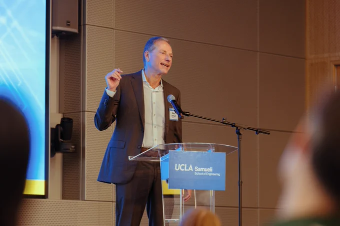 Henry Samueli stands at a podium with a microphone, speaking to an audience. The podium features a sign that reads "UCLA Samueli School of Engineering." The background includes a large screen displaying abstract blue graphics.