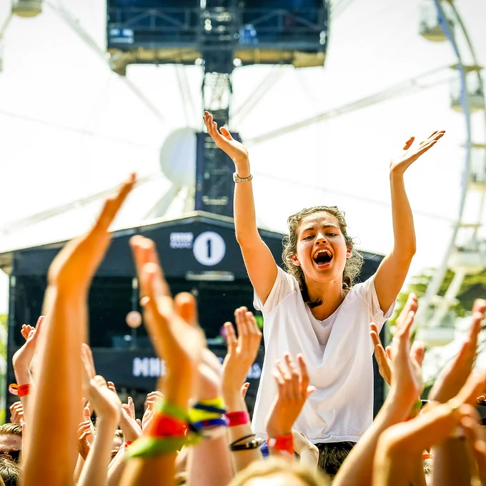Festival attendee in a white T-shirt sits above the crowd with both arms raised, smiling and cheering while surrounded by hands reaching upward.