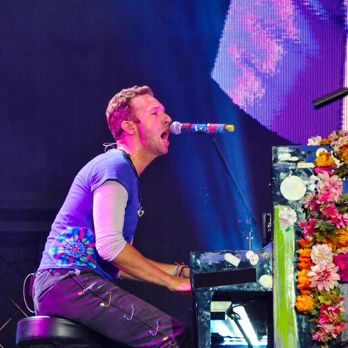 Musician sings into a microphone while playing a piano decorated with colorful flowers. Stage lighting casts pink and purple tones across the performance.