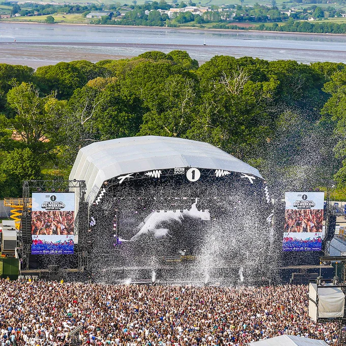 Wide view of a packed festival audience facing a large outdoor stage. White confetti bursts above the crowd, with trees and water visible in the background.