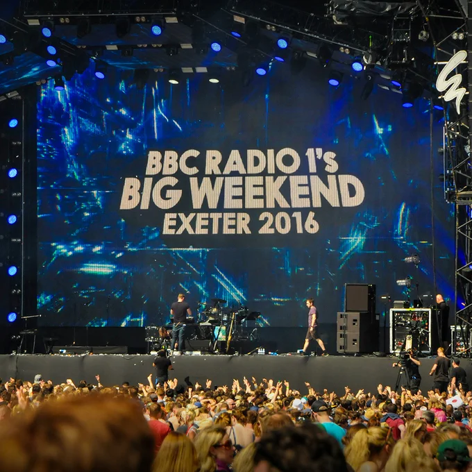 Crowd gathered in front of a large festival stage displaying the text “BBC Radio 1’s Big Weekend Exeter 2016.” Crew members and equipment are visible on stage between performances.