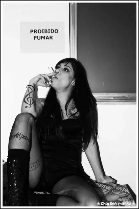 Foto em preto e branco de 2008. Pitty fuma sentada em frente a um aviso de Proibido Fumar. Foto de Charlie Messa.