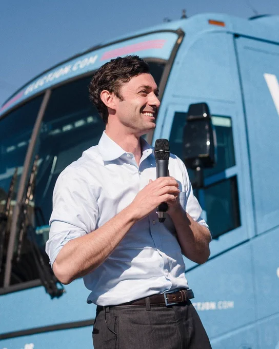 An image of Senator Jon Ossoff standing in front of a campaign bus. He is smiling with a microphone in his hand. 