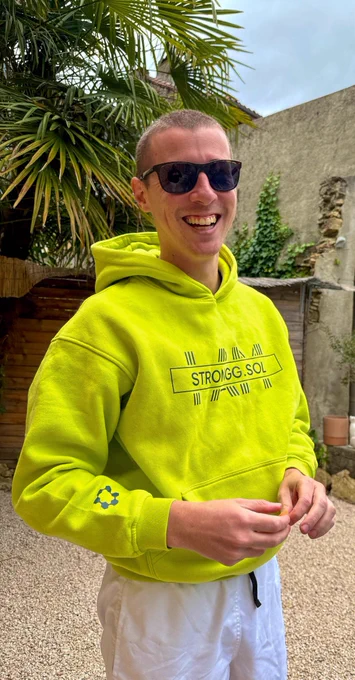 A smiling person in a bright yellow hoodie stands in a garden surrounded by palm leaves, exuding a cheerful vibe.