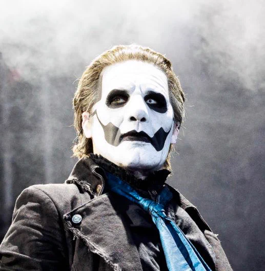 Picture of Papa Emeritus 4 from the band Ghost taken during a concert. It shows him from the chest up, body turned slightly to the right while his head is facing the viewer. His eyes are looking up to the upper left. He’s wearing a black jacket over a black shirt and blue necktie. The background is full of fog.