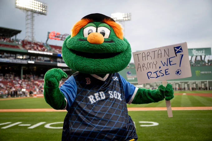 Wally the Green Monster stands on field at Fenway Park holding a sign that reads "Tartan Army, We Rise!" He is wearing the Scottish Celebration Night Red Sox Jersey.