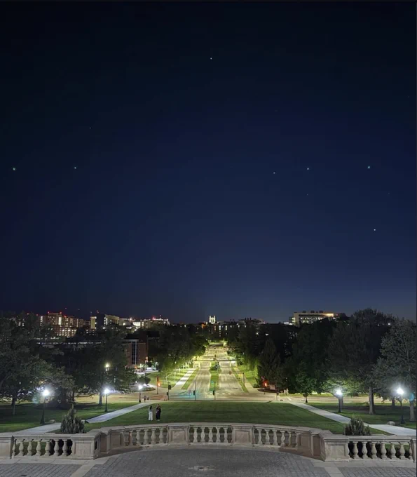 A nighttime Iowa city view of Iowa Ave, lined with trees and street lamps, leading towards campus buildings under a clear, starry sky.