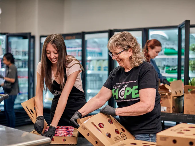 Two people are packing boxes of food in a food bank or grocery setting. One wears a shirt with "Feeding America" and "Hope" prominently displayed. They are surrounded by refrigerators stocked with various items. Everyone appears to be focused and engaged in their tasks.