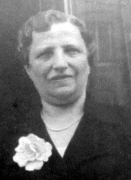 A black and white photograph of a woman wearing a dark blouse adorned with a large flower brooch, smiling and posing for the camera.
