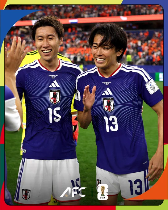 Two Japanese soccer players in blue jerseys display happiness and camaraderie after a match, with a blurred stadium background.