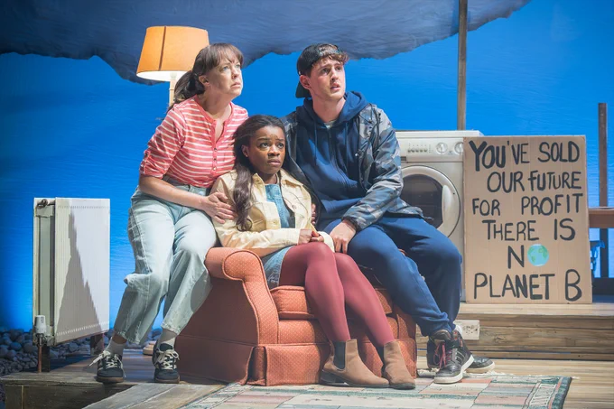 Three actors huddled together on a chair on stage, surrounded by a radiator, lamp, washing machine and protest sign