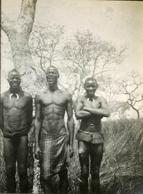 Photograph (black and white), from an album: view three adult males standing side by side and facing the camera. The man on the left appears to be smiling/laughing and has his arms positioned behind his back, he is also wearing a personal ornament around his neck in the shape of a crescent and a waisted wrapper. The man in the middle is wearing a long cloth wrap around his waist with his arms at either side. On the right is a shorter man with his arms crossed in front of his body, also wearing a crescent-like personal ornament around his neck. In the distance are several tall trees. Southern Nigeria.
Gelatin silver print