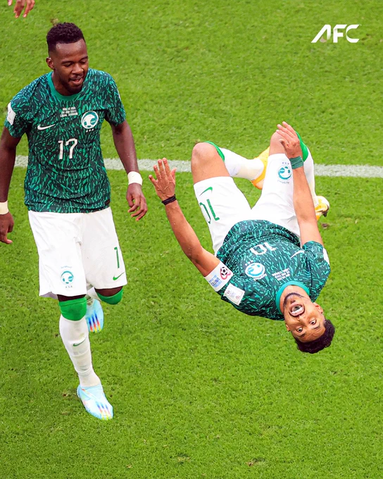 A soccer player performs an acrobatic flip in celebration on the field, while a teammate looks on with excitement. The mood is energetic and celebratory.