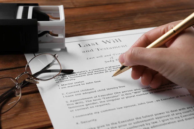 Person signing a Last Will and Testament document with a gold pen on a wooden table beside reading glasses and a black stamp holder.