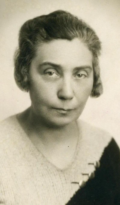 Black and white portrait of a woman with a serious expression, wearing a knitted sweater with a distinct geometric pattern on the collar. The person has short hair combed back and is looking directly at the camera.
