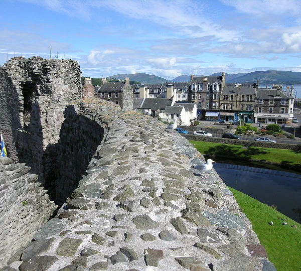 Today's 'Where in Scotland' image. It is a squarish image showing the top of a thick stone wall entering the bottom of the frame and curving to the left where it meets part of a ruined structure in the upper left of the frame. To the right and beyond the curving wall is a view down to grass and part of a curving water feature, with part of a town in the background and sea and hills beyond them. The scene is in sunlight.