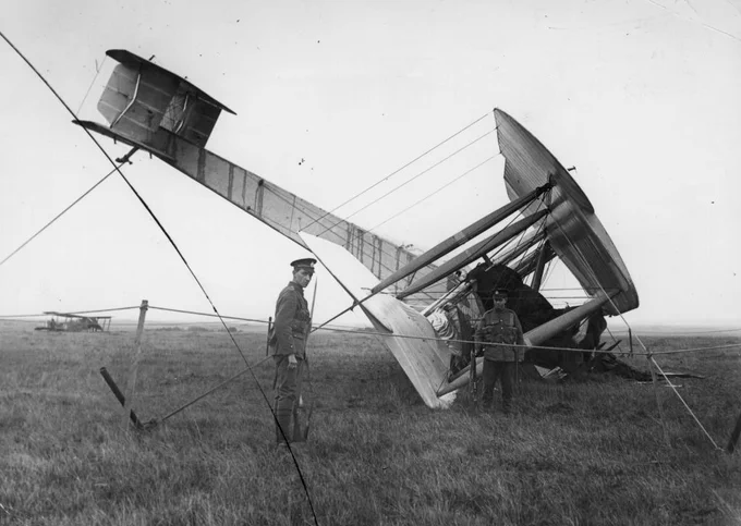 A biplane lies crashed in a field with two individuals standing nearby observing the wreckage. The aircraft's wings and tail show significant damage. Another biplane is visible in the distance on the grassy landscape.